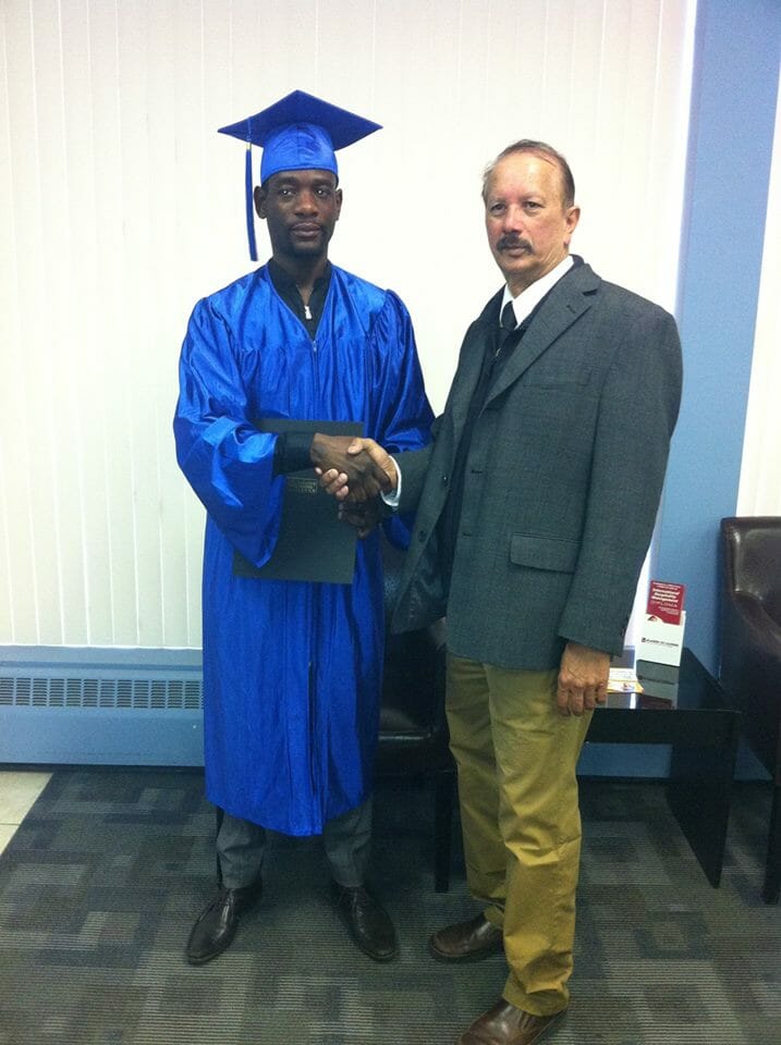 Graduate in blue cap and gown shaking hands during a certificate presentation ceremony.