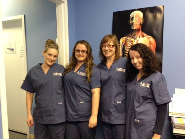 Group of healthcare students wearing scrubs standing in a classroom with a human anatomy poster in the background.