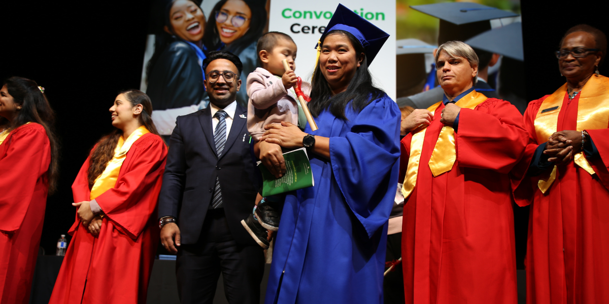 A graduate in blue cap and gown holding her child during an Academy of Learning Career College convocation ceremony, surrounded by faculty in red robes with gold stoles. This milestone represents the career advancement possible with proper financial planning and student loan management.