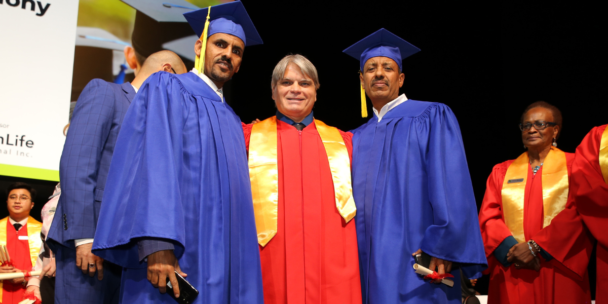 Two male graduates in blue regalia standing with an instructor in red academic robes at an Academy of Learning Career College convocation ceremony. Many AOLCC graduates successfully utilize income-driven repayment options in Canada to manage education costs while building their careers.