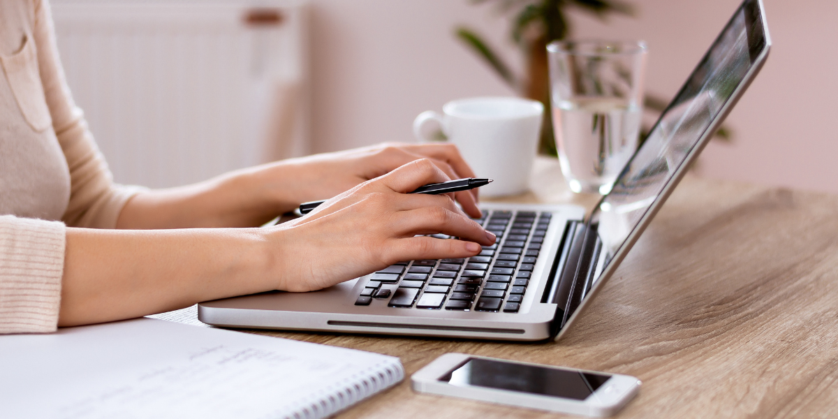 Close-up view of hands typing on laptop keyboard with notebook and smartphone nearby, illustrating how Microsoft Office applications enhance productivity in comfortable work settings.